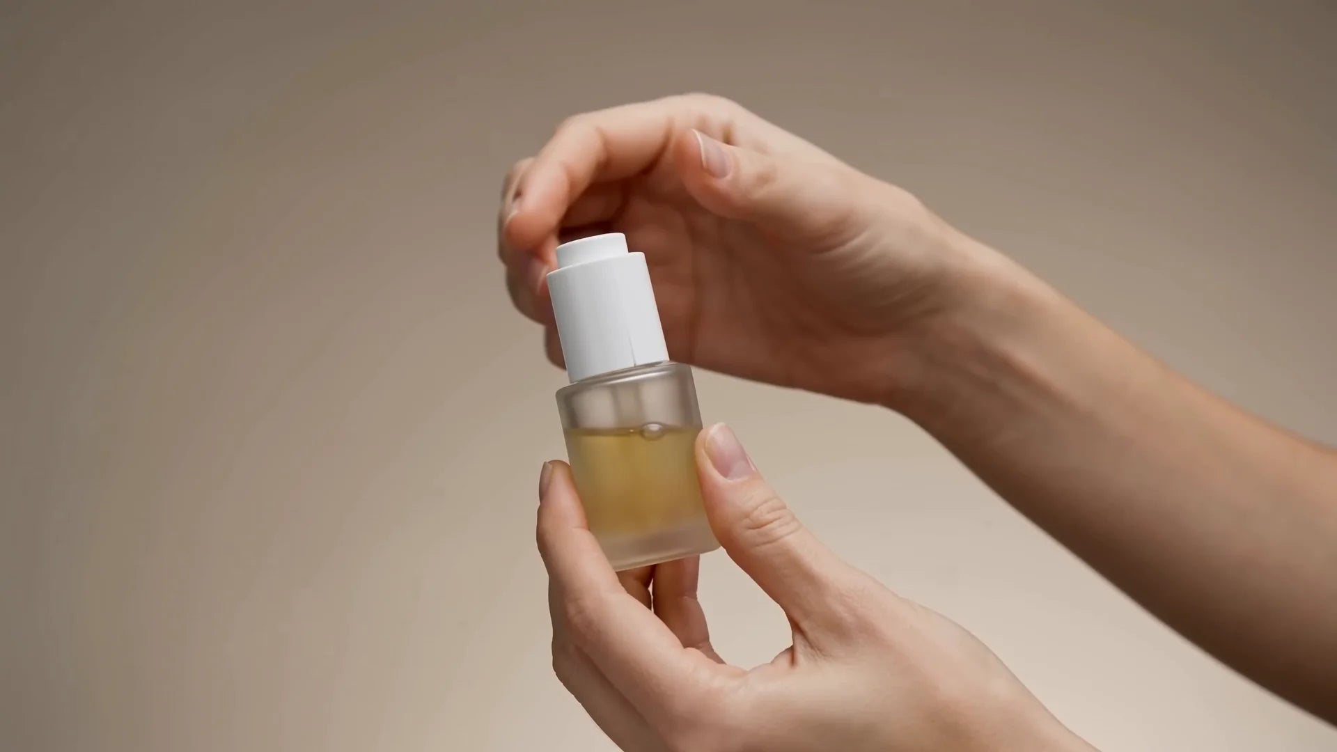 Des mains ouvrent un flacon de sérum au liquide doré, sur fond écru.